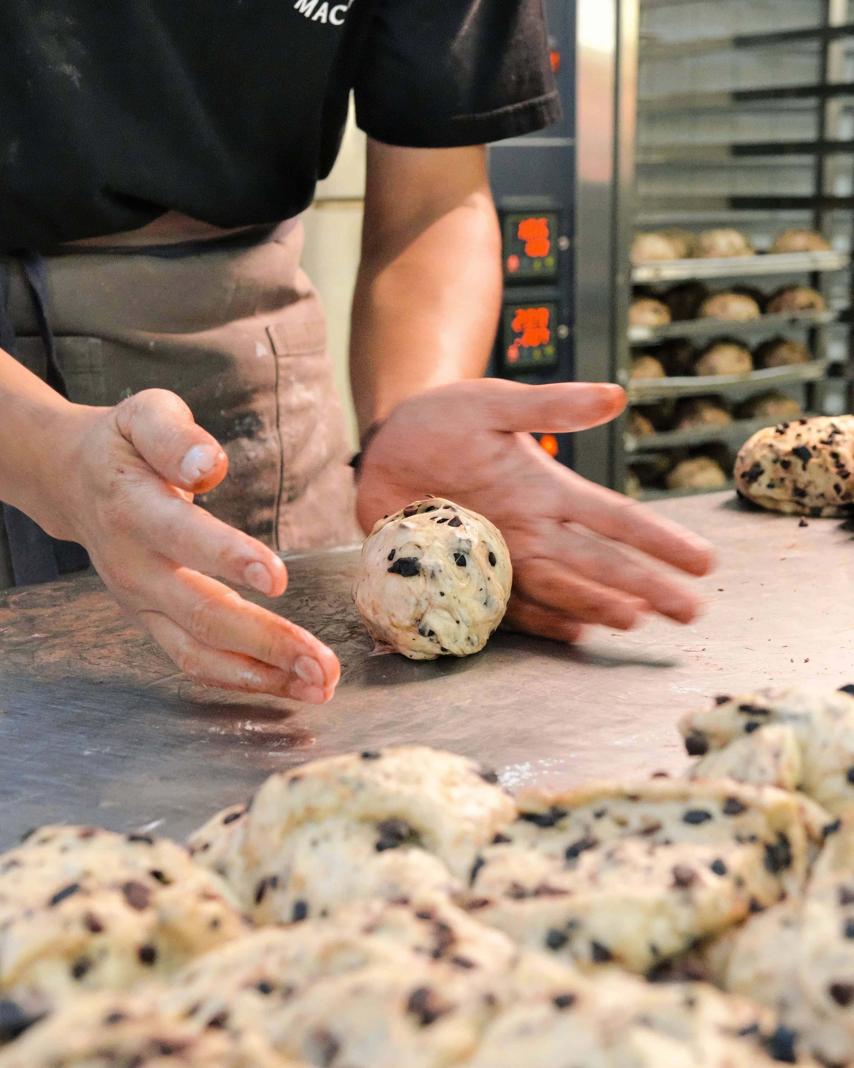 Two hands rolling a round of chocolate chip dough on the steel bench