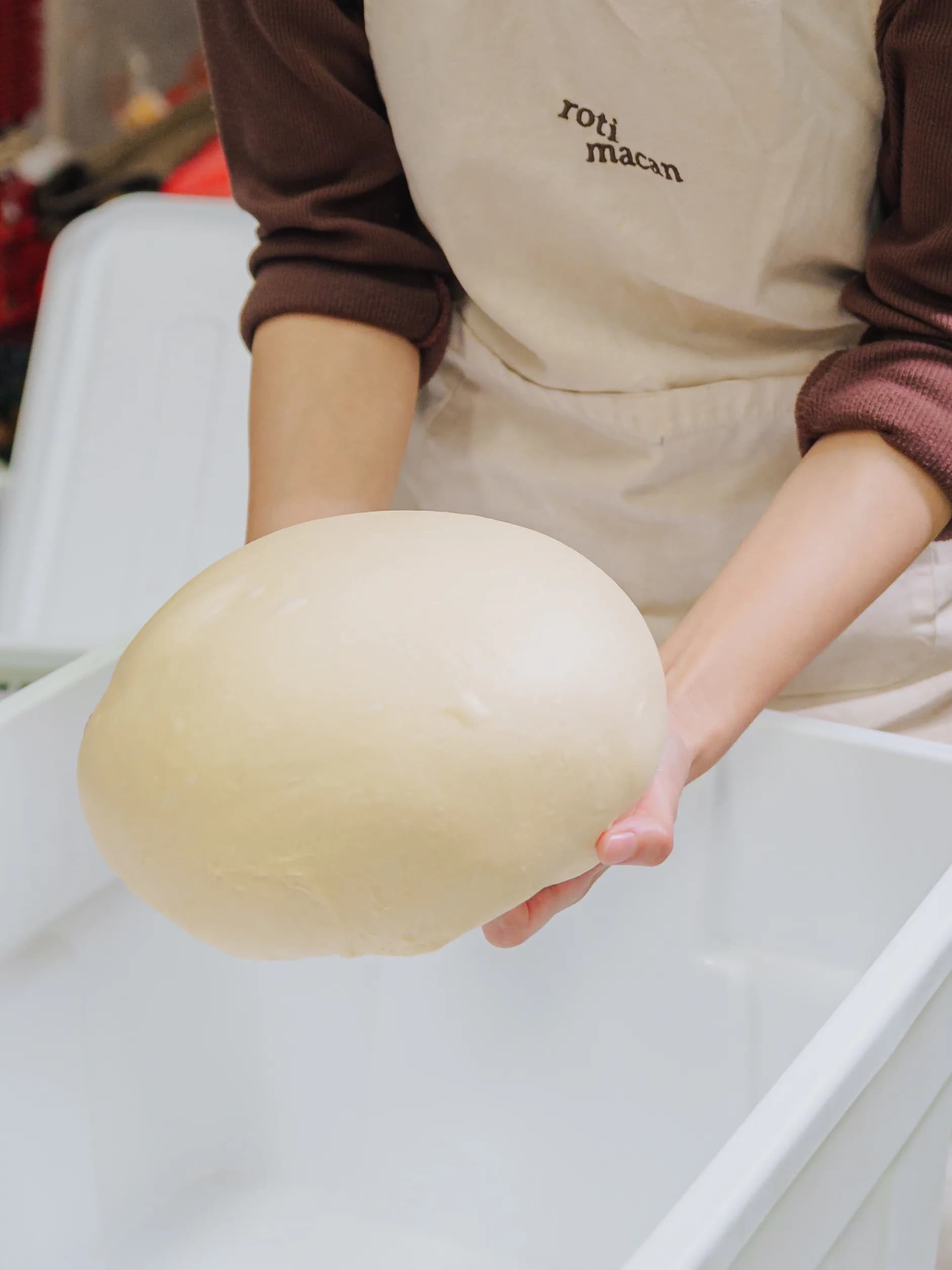 A baker in a roti macan apron lifting a round of proofed dough out of the tub